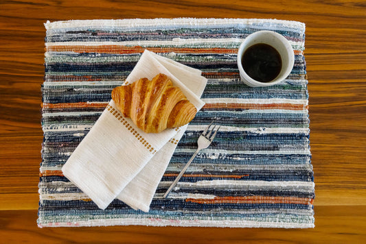 Adesé placemat with a croissant, coffee, and fork on a wooden surface