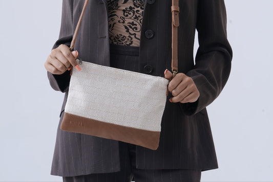 A model wearing a dark gray pinstripe blazer and black lace top holds a rectangular chiban bag with a white linen-textured body and brown leather trim and strap against a light gray studio background.