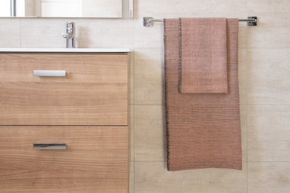 Handwoven bath towel in blush, displayed in a bathroom setting, with a wooden cabinet and towel rack.
